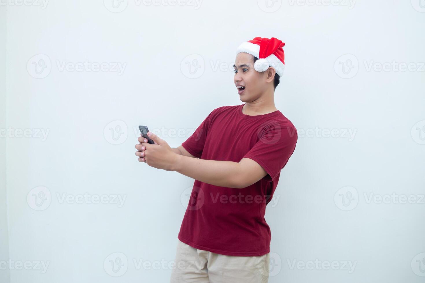Young Asian man wearing a Santa Claus hat holding a smartphone and expressing a smile, shock and surprise isolated by a white background for visual communication photo