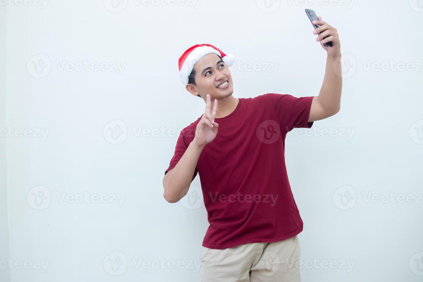 joven asiático hombre vistiendo un Papa Noel claus sombrero, participación un teléfono inteligente y tomando un selfie aislado en contra un blanco antecedentes para visual comunicación foto