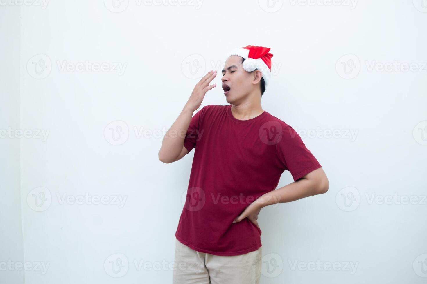 joven asiático hombre vistiendo un Papa Noel claus sombrero expresando cansancio y bostezando aislado por un blanco antecedentes para visual comunicación foto