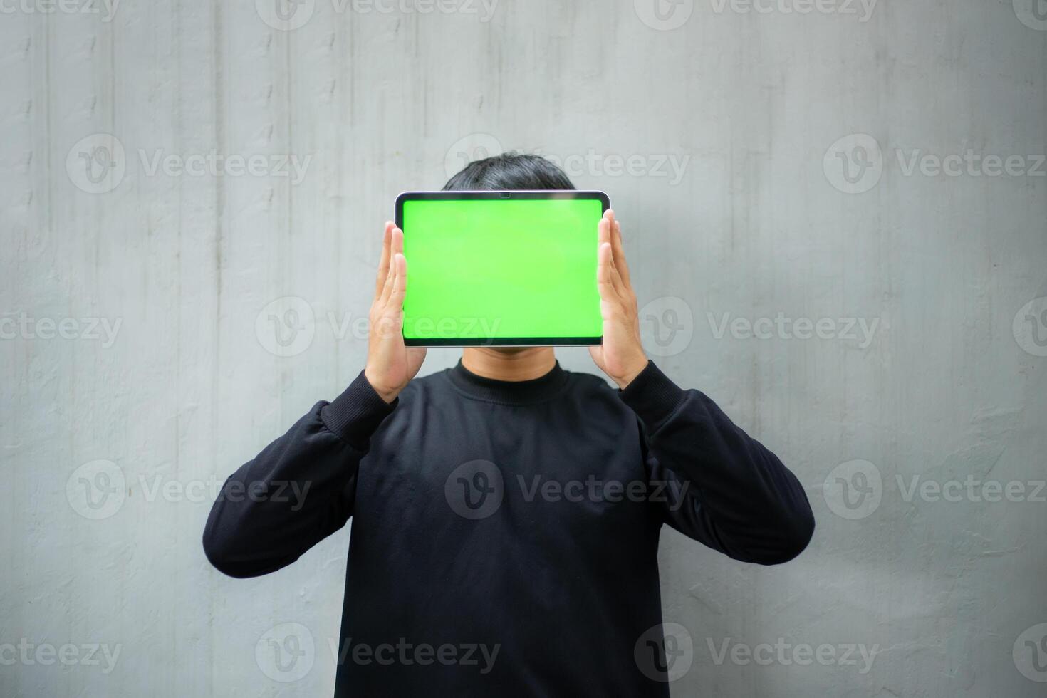 Young Asian man with a black sweater holding and pointing at an iPad tablet green screen mockup photo