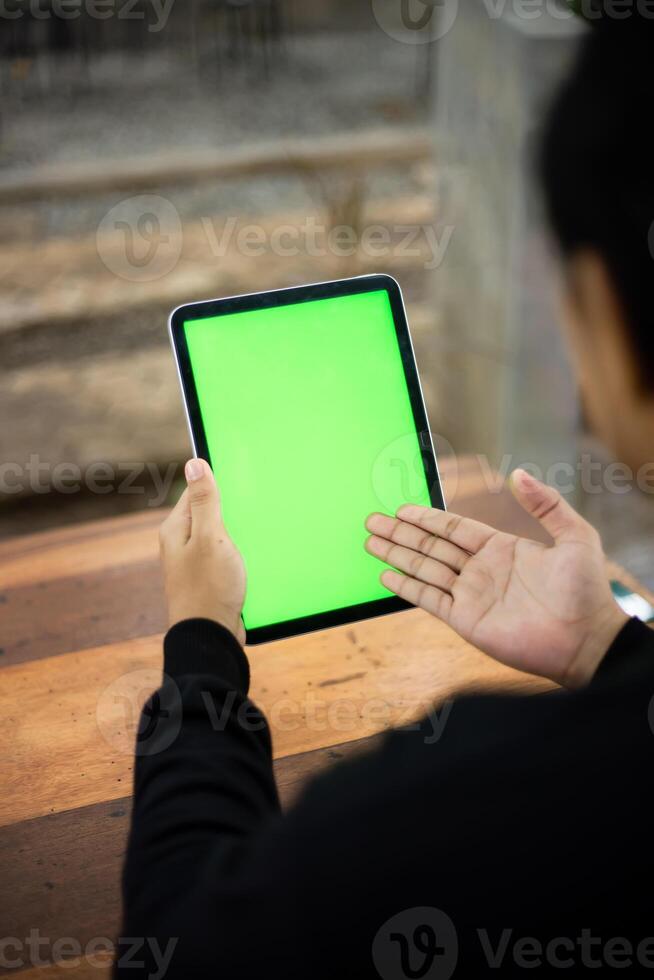 Mock up photo of a close up shot featuring a mans hand holding an iPad tablet with a green screen against the background of a wood cafe table