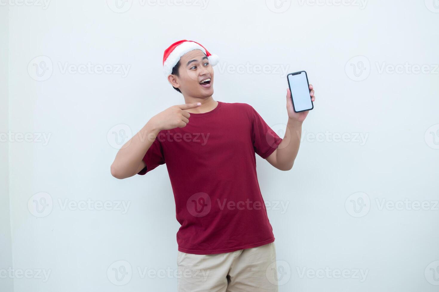 Young Asian man wearing a Santa Claus hat holding a smartphone, and pointing to the phone with a smile, look of shock, and surprise isolated against a white background for visual communication photo