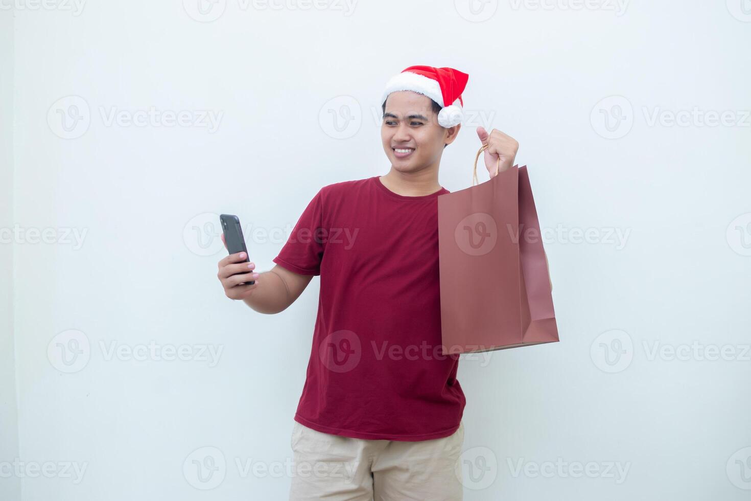 joven asiático hombre vistiendo un Papa Noel claus sombrero participación un teléfono inteligente y un compras bolso con expresiones de sonrisa, choque, y sorpresa, aislado en contra un blanco antecedentes para visual comunicación foto