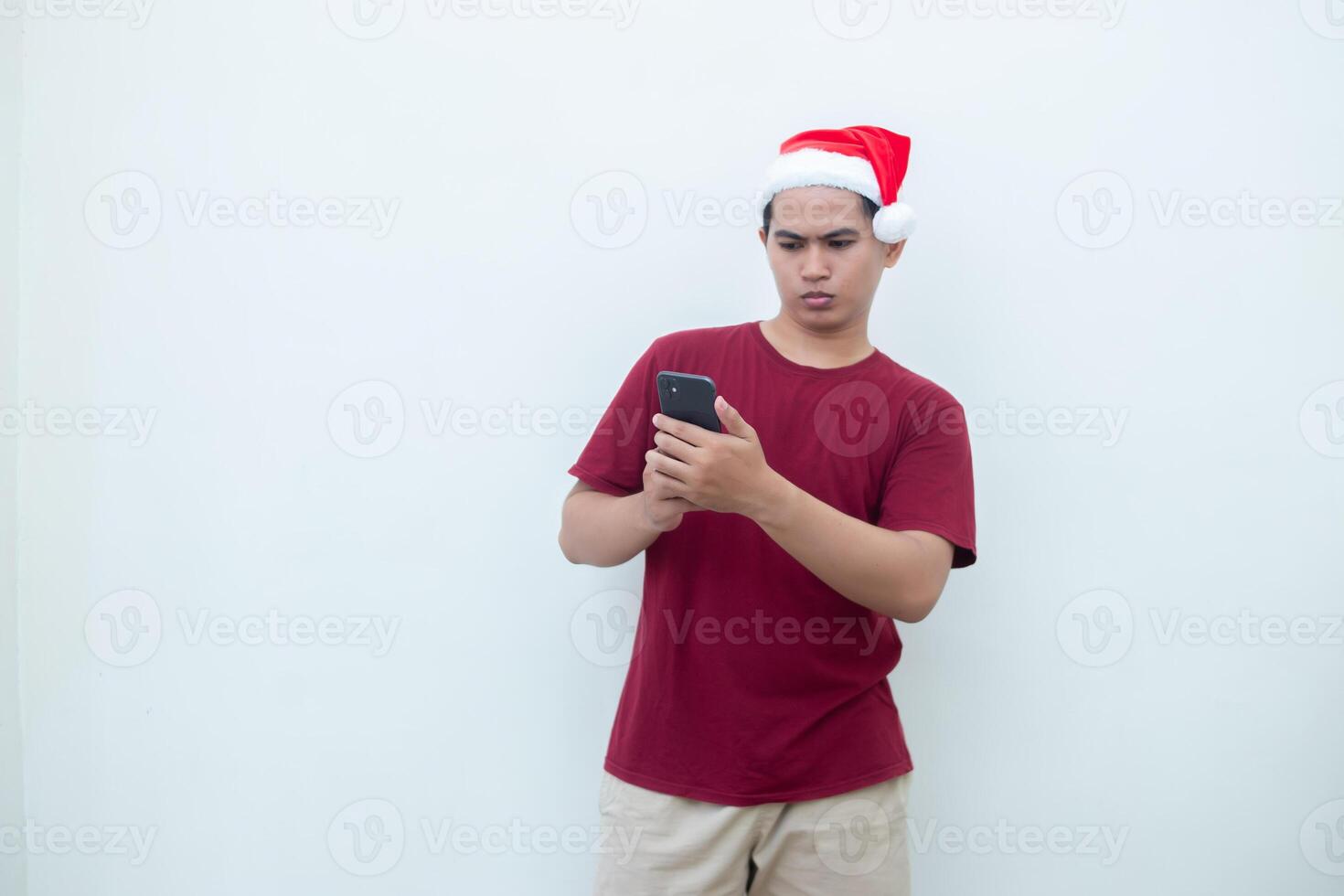 Young Asian man wearing a Santa Claus hat holding a smartphone and expressing a smile, shock and surprise isolated by a white background for visual communication photo