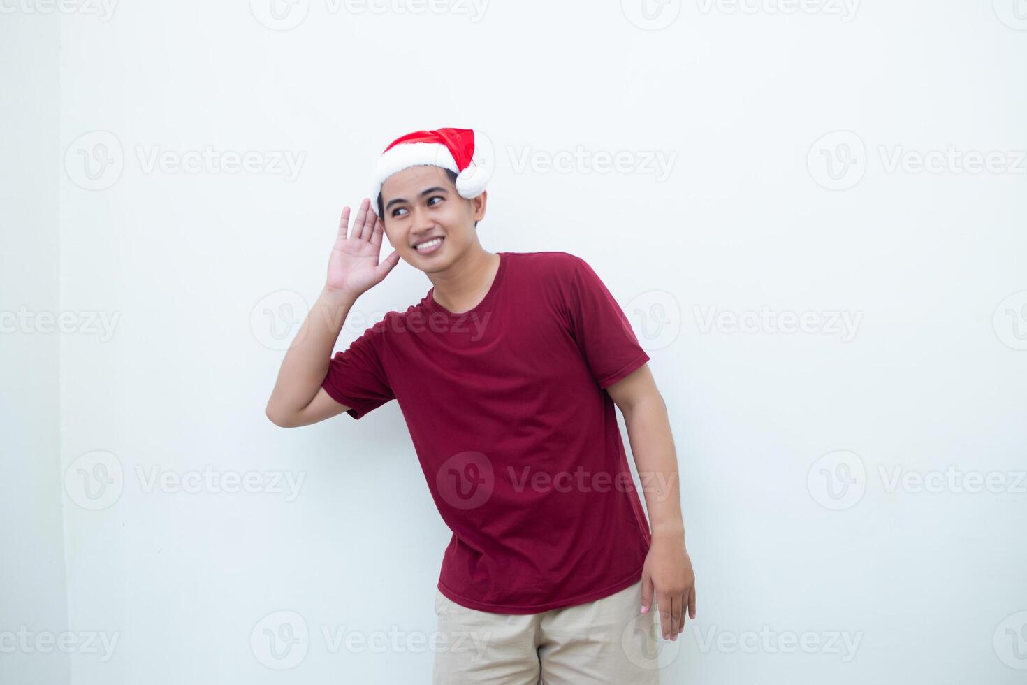 Young Asian man wearing a Santa Claus hat expressing listening with hand to ear isolated by a white background for visual communication photo