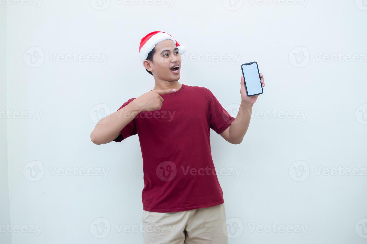 Young Asian man wearing a Santa Claus hat holding a smartphone, and pointing to the phone with a smile, look of shock, and surprise isolated against a white background for visual communication photo