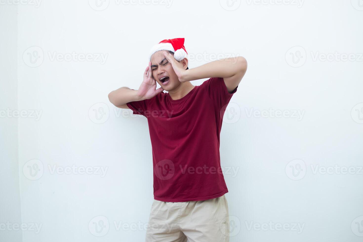 Young Asian man wearing a Santa Claus hat expressing a headache by holding his head and showing signs of pain isolated by a white background for visual communication photo