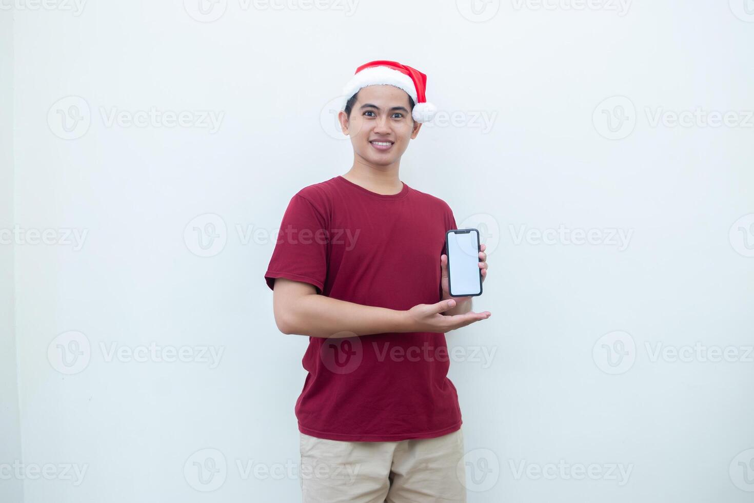 joven asiático hombre vistiendo un Papa Noel claus sombrero participación un teléfono inteligente, y señalando a el teléfono con un sonrisa, Mira de choque, y sorpresa aislado en contra un blanco antecedentes para visual comunicación foto