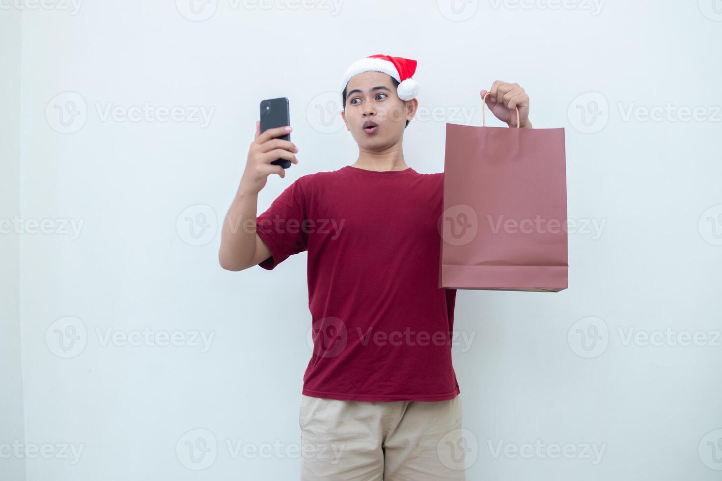 joven asiático hombre vistiendo un Papa Noel claus sombrero participación un teléfono inteligente y un compras bolso con expresiones de sonrisa, choque, y sorpresa, aislado en contra un blanco antecedentes para visual comunicación foto