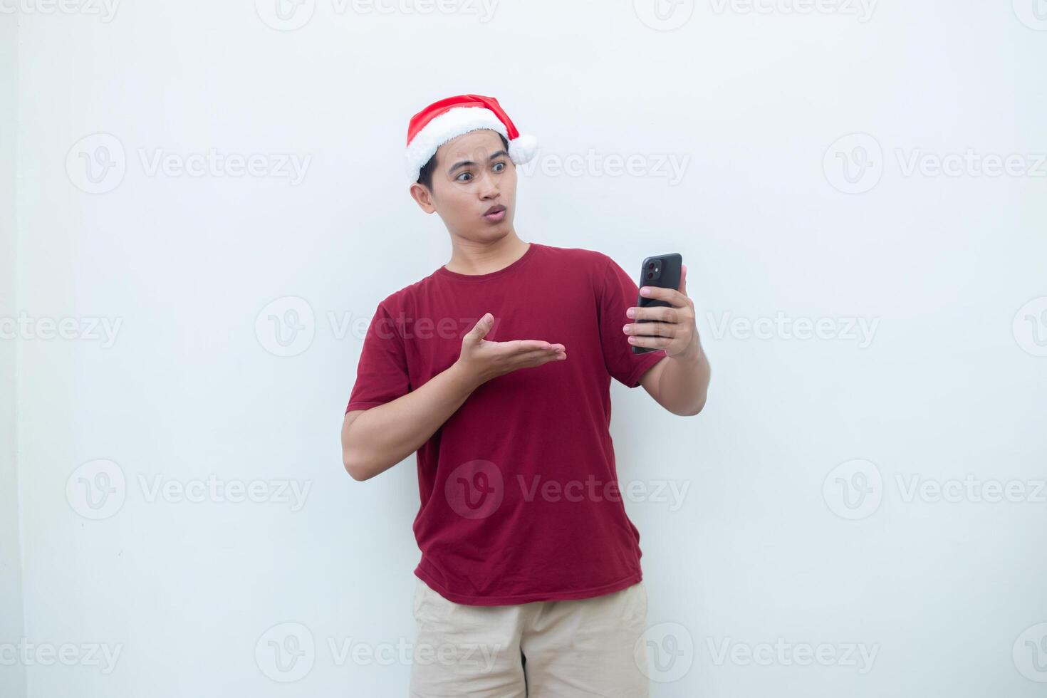 Young Asian man wearing a Santa Claus hat holding a smartphone and expressing a smile, surprise, and pointing to the side, isolated by a white background for visual communication photo