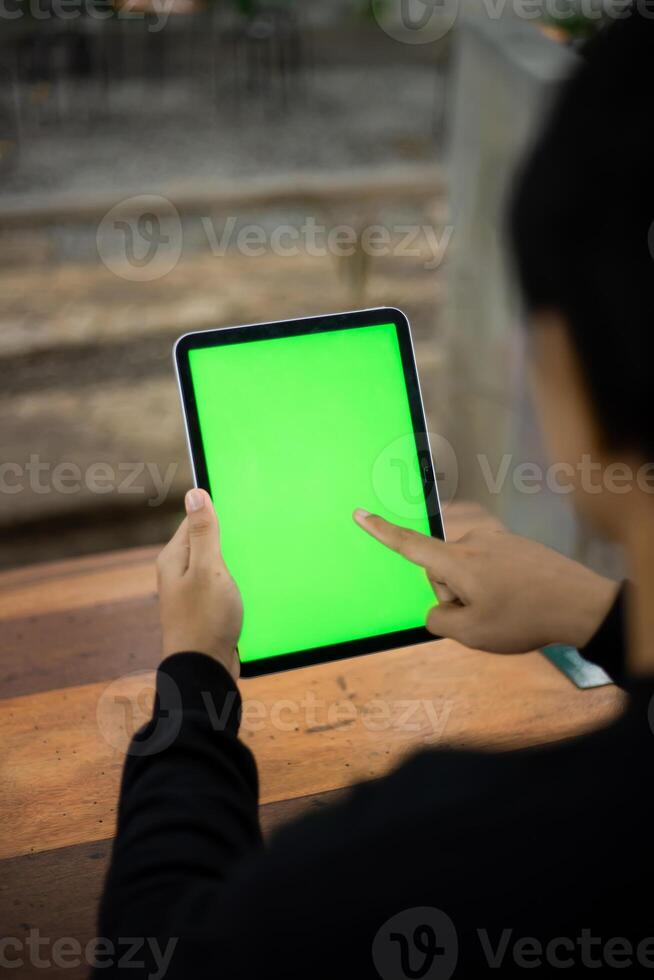 Mock up photo of a close up shot featuring a mans hand holding an iPad tablet with a green screen against the background of a wood cafe table