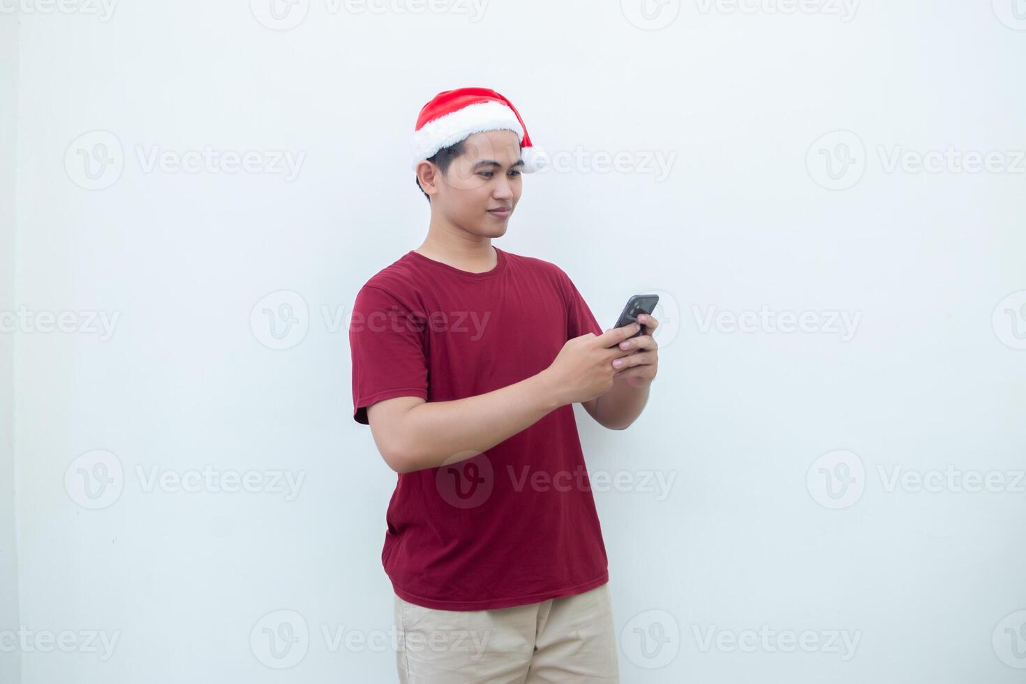Young Asian man wearing a Santa Claus hat holding a smartphone and expressing a smile, shock and surprise isolated by a white background for visual communication photo