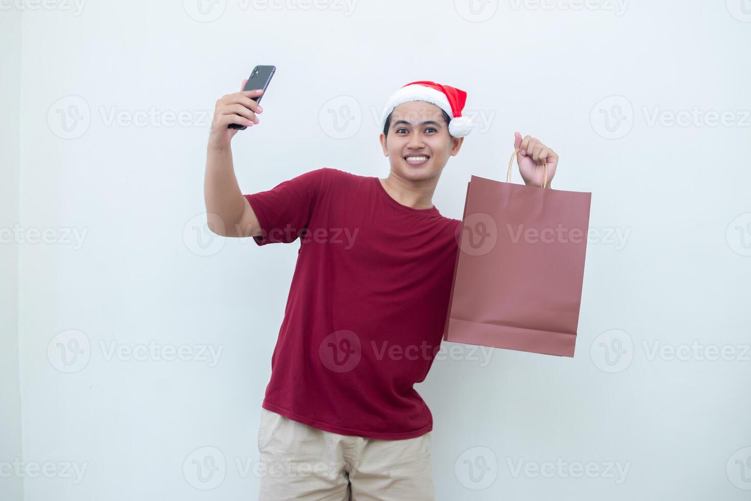 joven asiático hombre vistiendo un Papa Noel claus sombrero participación un teléfono inteligente y un compras bolso con expresiones de sonrisa, choque, y sorpresa, aislado en contra un blanco antecedentes para visual comunicación foto