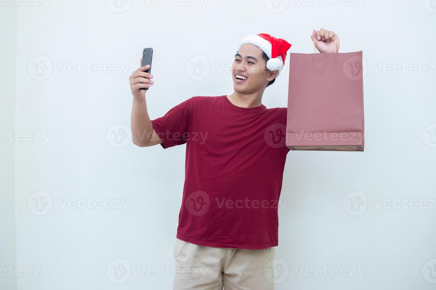 Young Asian man wearing a Santa Claus hat holding a smartphone and a shopping bag with expressions of smile, shock, and surprise, isolated against a white background for visual communication photo