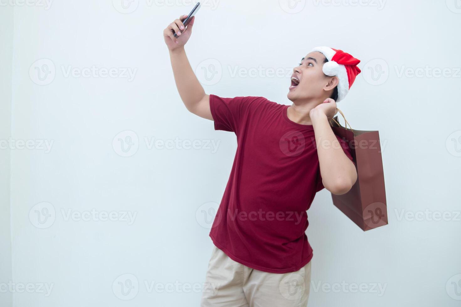 Young Asian man wearing a Santa Claus hat holding a smartphone and a shopping bag with expressions of smile, shock, and surprise, isolated against a white background for visual communication photo