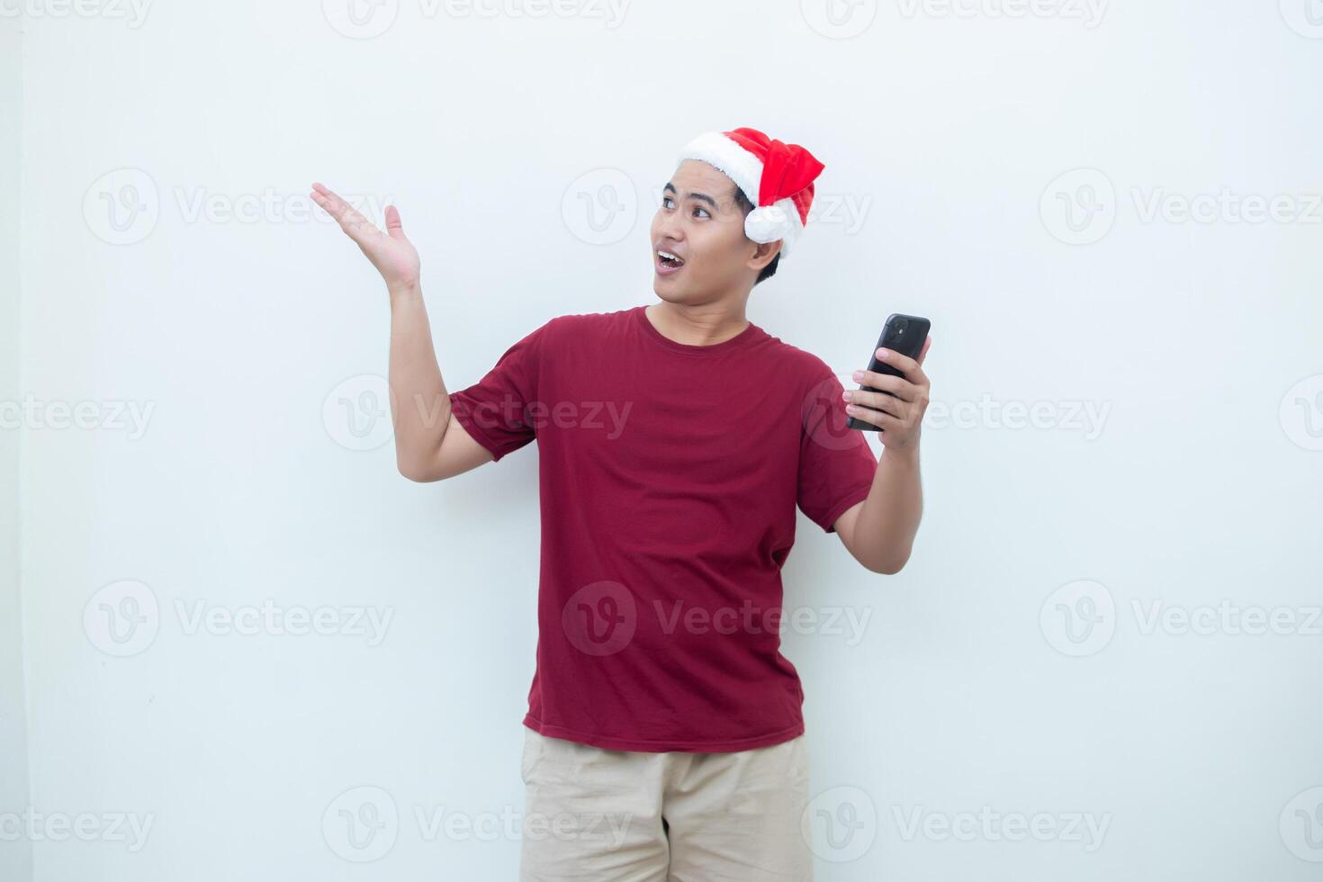 Young Asian man wearing a Santa Claus hat holding a smartphone and expressing a smile, surprise, and pointing to the side, isolated by a white background for visual communication photo