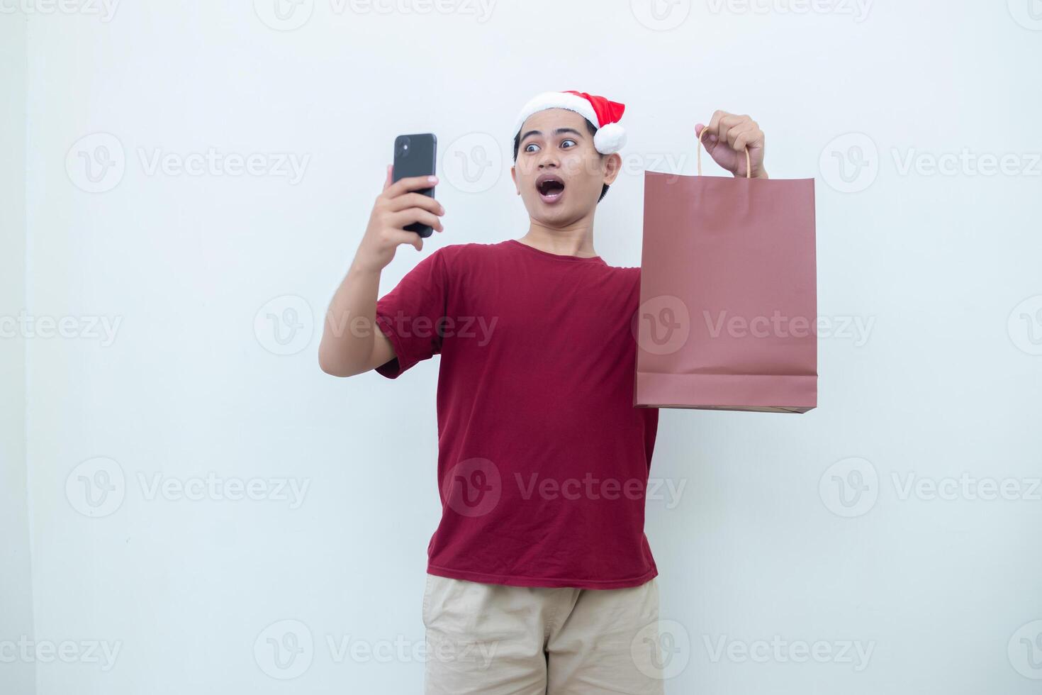 Young Asian man wearing a Santa Claus hat holding a smartphone and a shopping bag with expressions of smile, shock, and surprise, isolated against a white background for visual communication photo