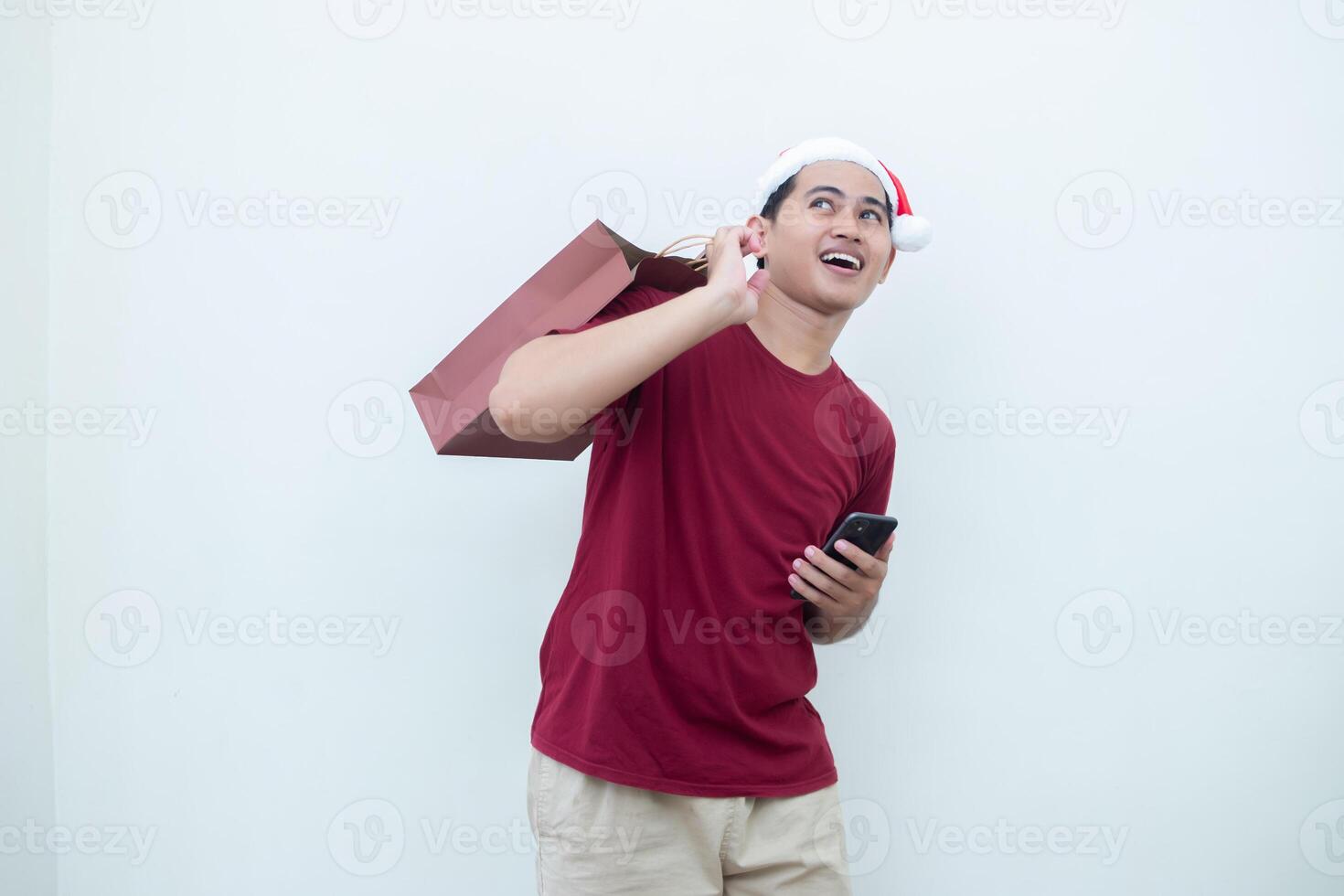 Young Asian man wearing a Santa Claus hat holding a smartphone and a shopping bag with expressions of smile, shock, and surprise, isolated against a white background for visual communication photo
