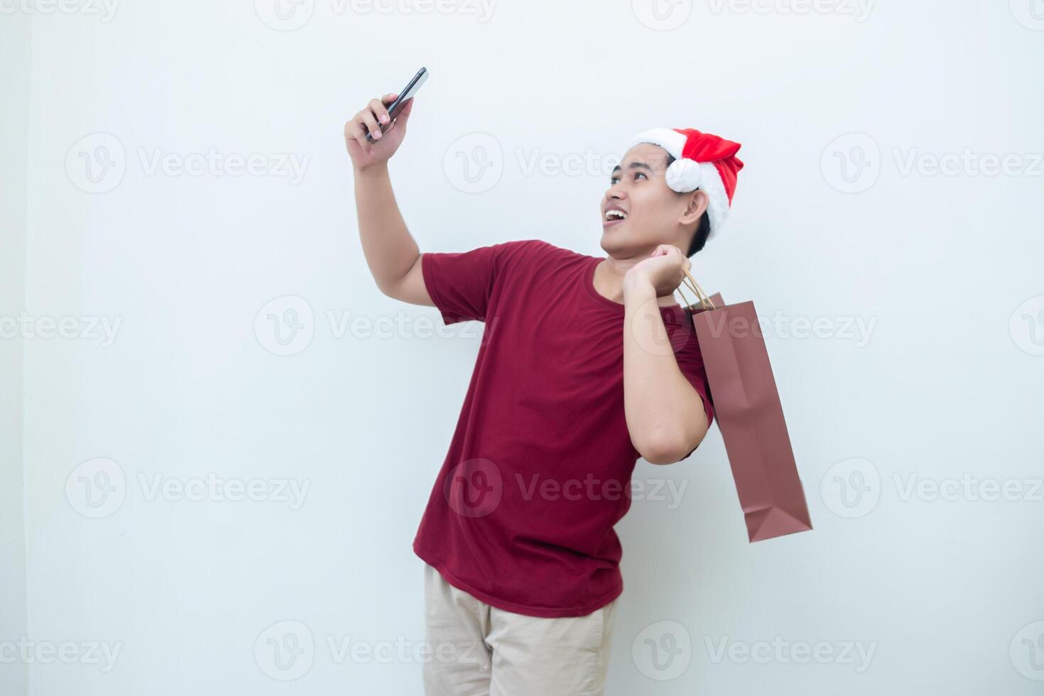 Young Asian man wearing a Santa Claus hat holding a smartphone and a shopping bag with expressions of smile, shock, and surprise, isolated against a white background for visual communication photo