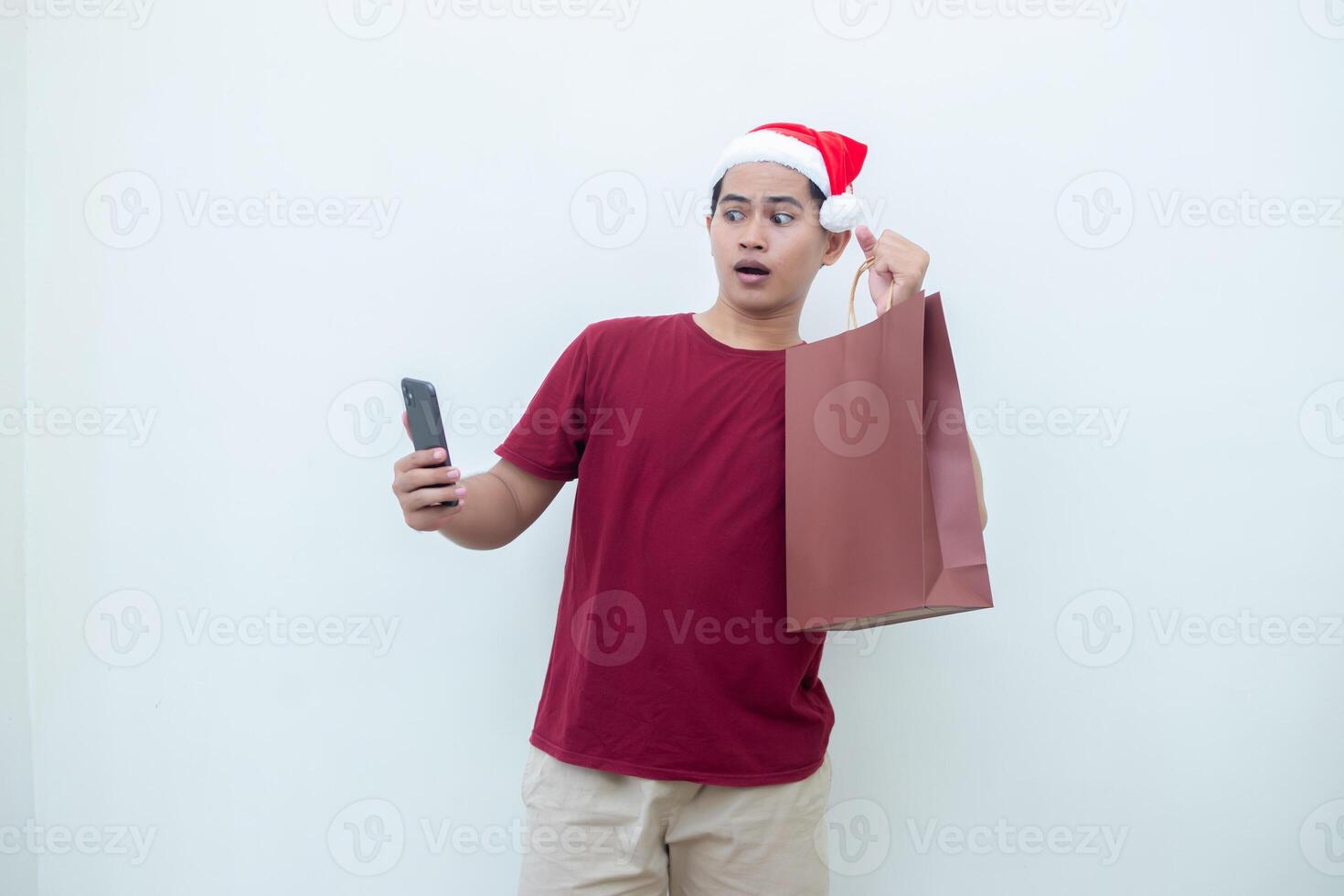 joven asiático hombre vistiendo un Papa Noel claus sombrero participación un teléfono inteligente y un compras bolso con expresiones de sonrisa, choque, y sorpresa, aislado en contra un blanco antecedentes para visual comunicación foto