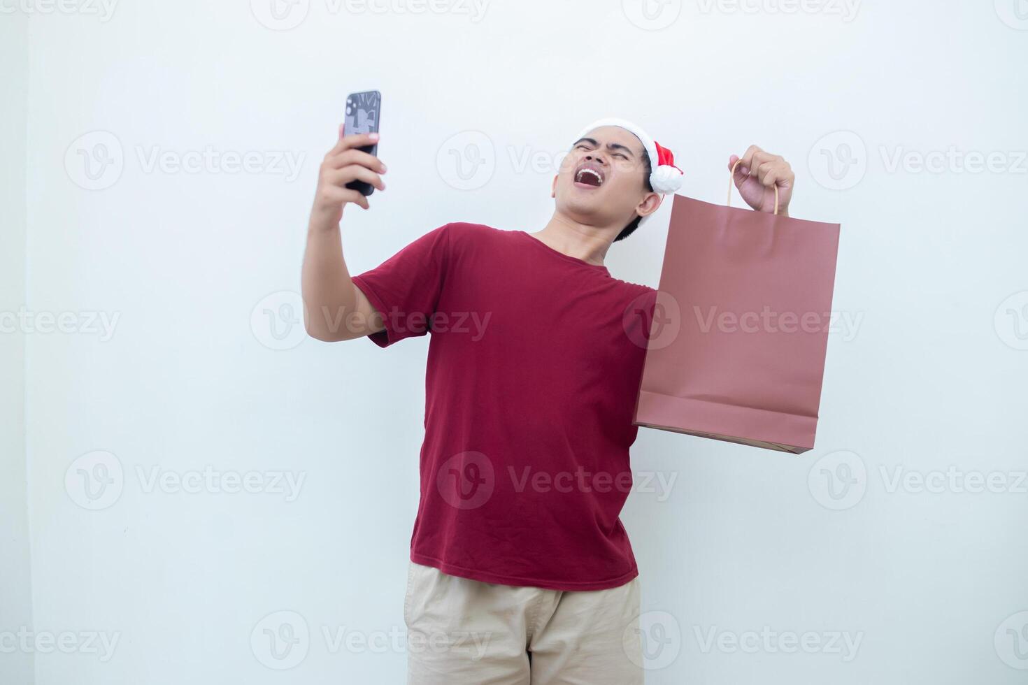 Young Asian man wearing a Santa Claus hat holding a smartphone and a shopping bag with expressions of smile, shock, and surprise, isolated against a white background for visual communication photo
