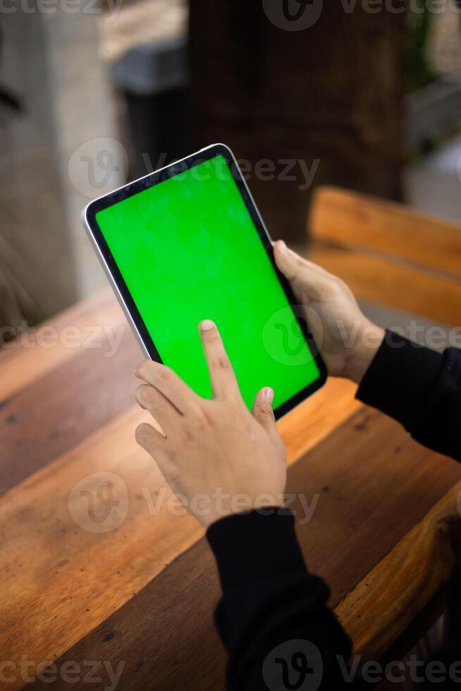 Mock up photo of a close up shot featuring a mans hand holding an iPad tablet with a green screen against the background of a wood cafe table