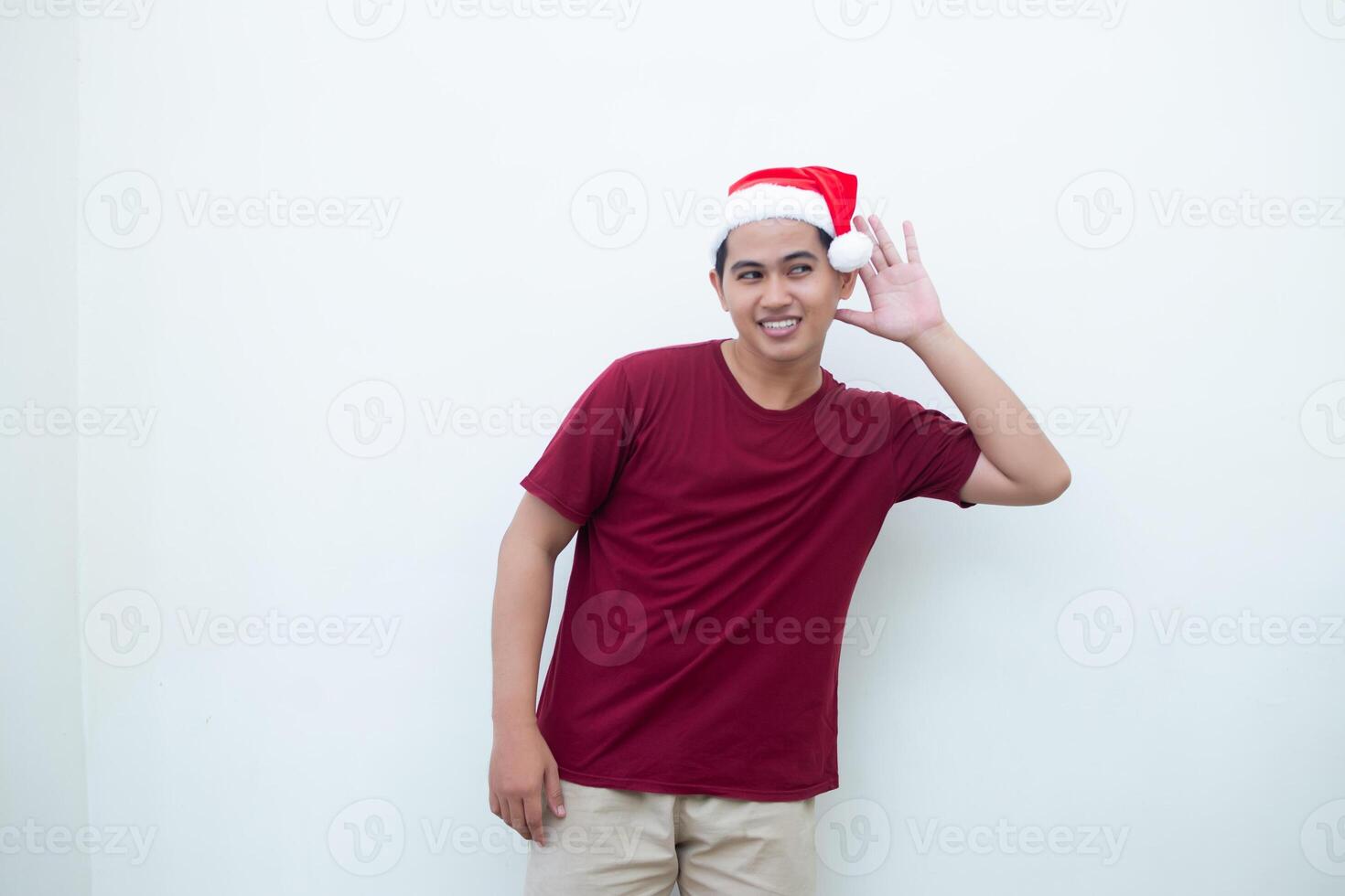 Young Asian man wearing a Santa Claus hat expressing listening with hand to ear isolated by a white background for visual communication photo