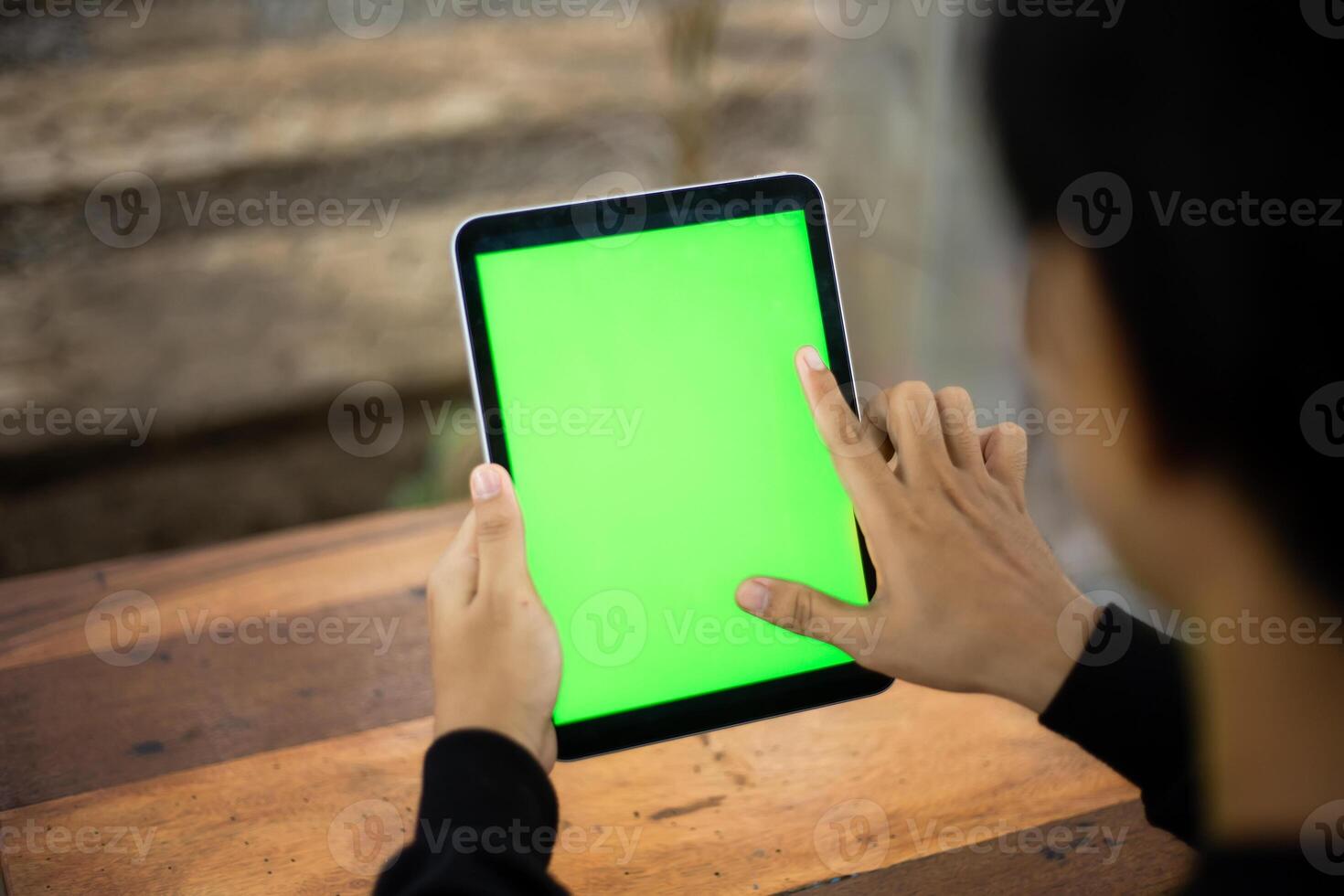 Mock up photo of a close up shot featuring a mans hand holding an iPad tablet with a green screen against the background of a wood cafe table