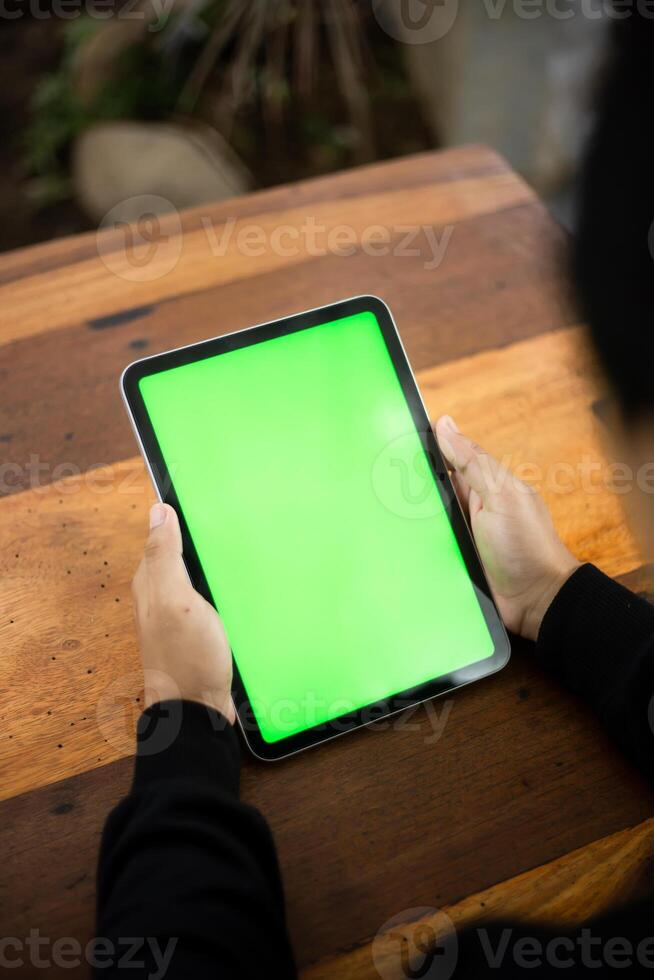 Mock up photo of a close up shot featuring a mans hand holding an iPad tablet with a green screen against the background of a wood cafe table