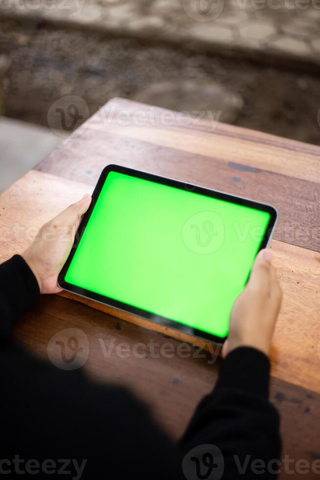 Mock up photo of a close up shot featuring a mans hand holding an iPad tablet with a green screen against the background of a wood cafe table