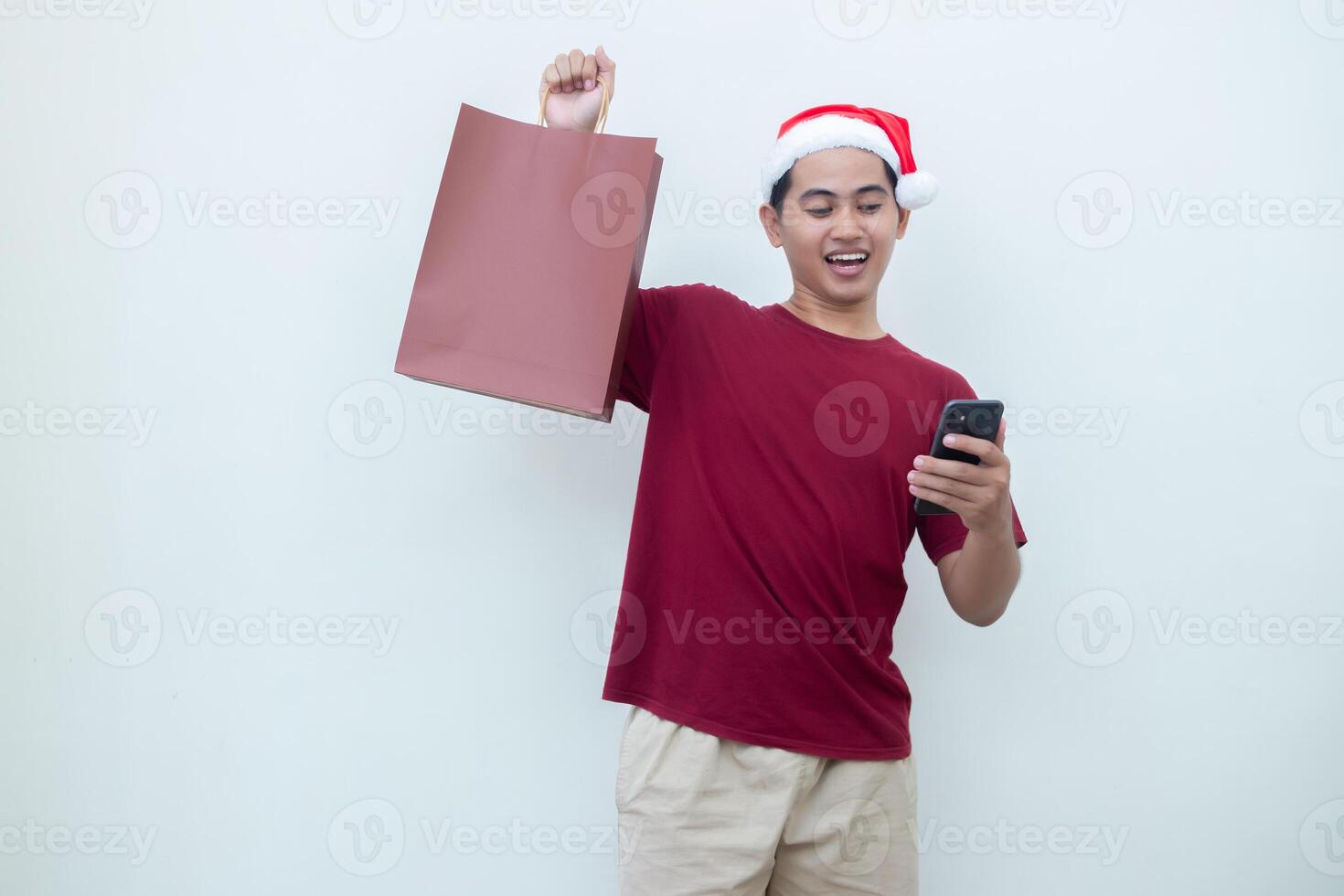 joven asiático hombre vistiendo un Papa Noel claus sombrero participación un teléfono inteligente y un compras bolso con expresiones de sonrisa, choque, y sorpresa, aislado en contra un blanco antecedentes para visual comunicación foto