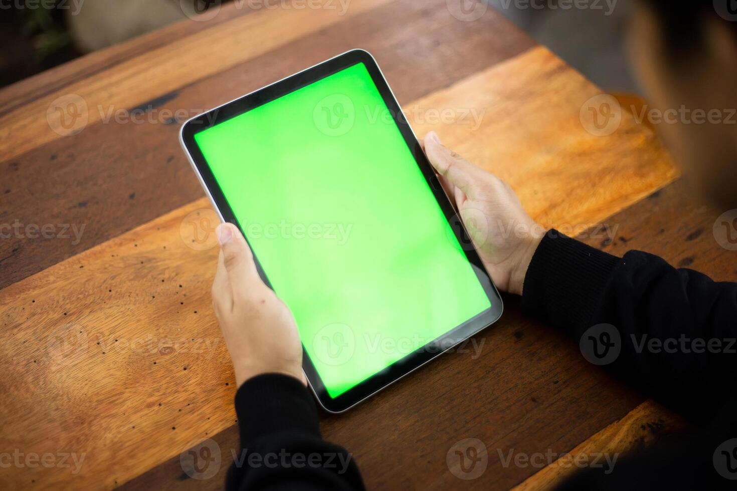 Mock up photo of a close up shot featuring a mans hand holding an iPad tablet with a green screen against the background of a wood cafe table