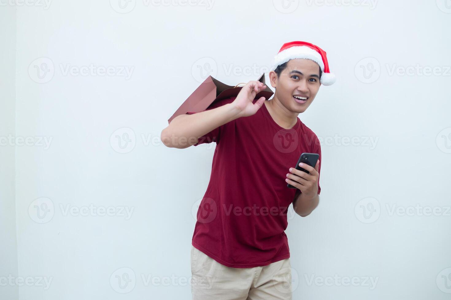 Young Asian man wearing a Santa Claus hat holding a smartphone and a shopping bag with expressions of smile, shock, and surprise, isolated against a white background for visual communication photo