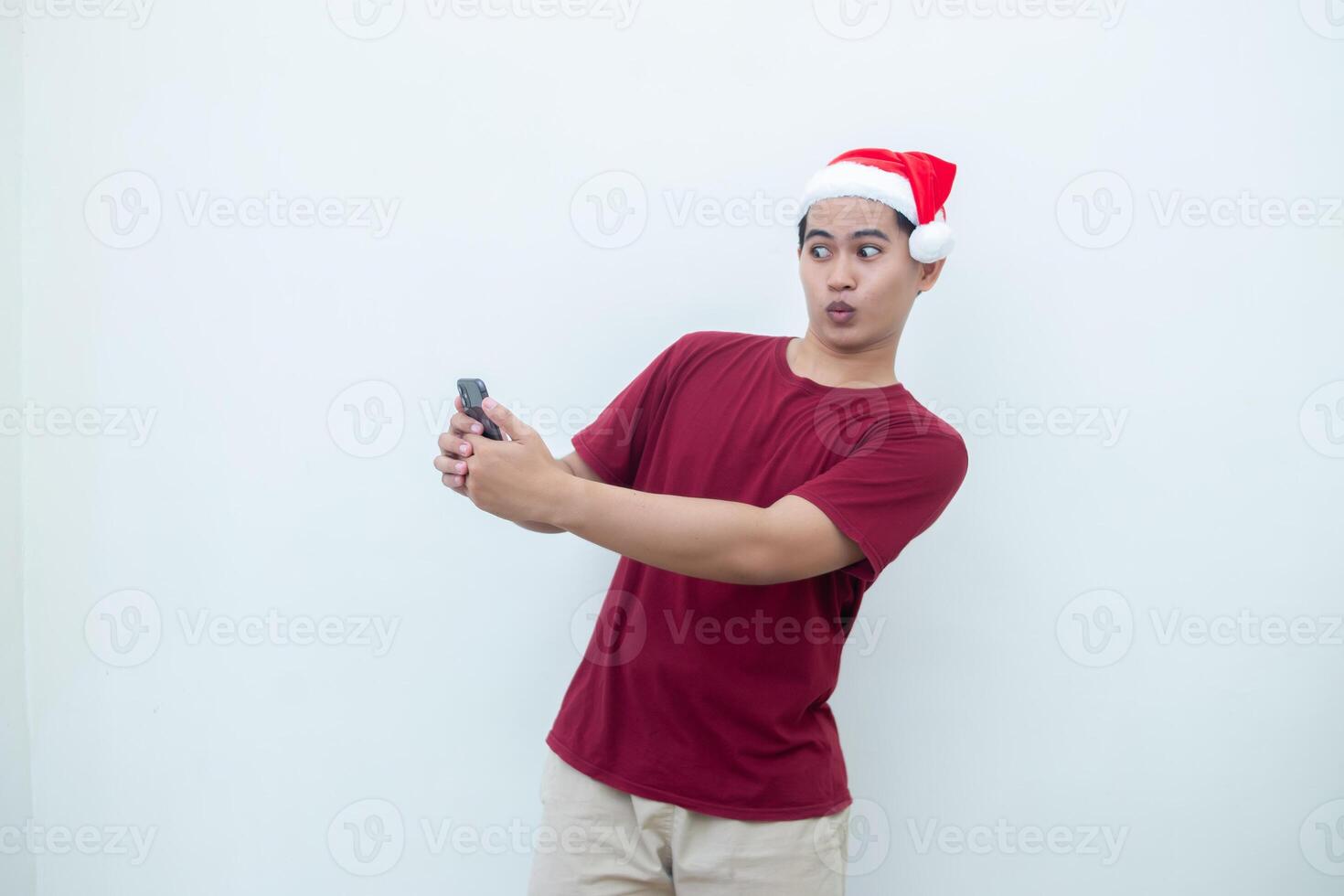 Young Asian man wearing a Santa Claus hat holding a smartphone and expressing a smile, shock and surprise isolated by a white background for visual communication photo