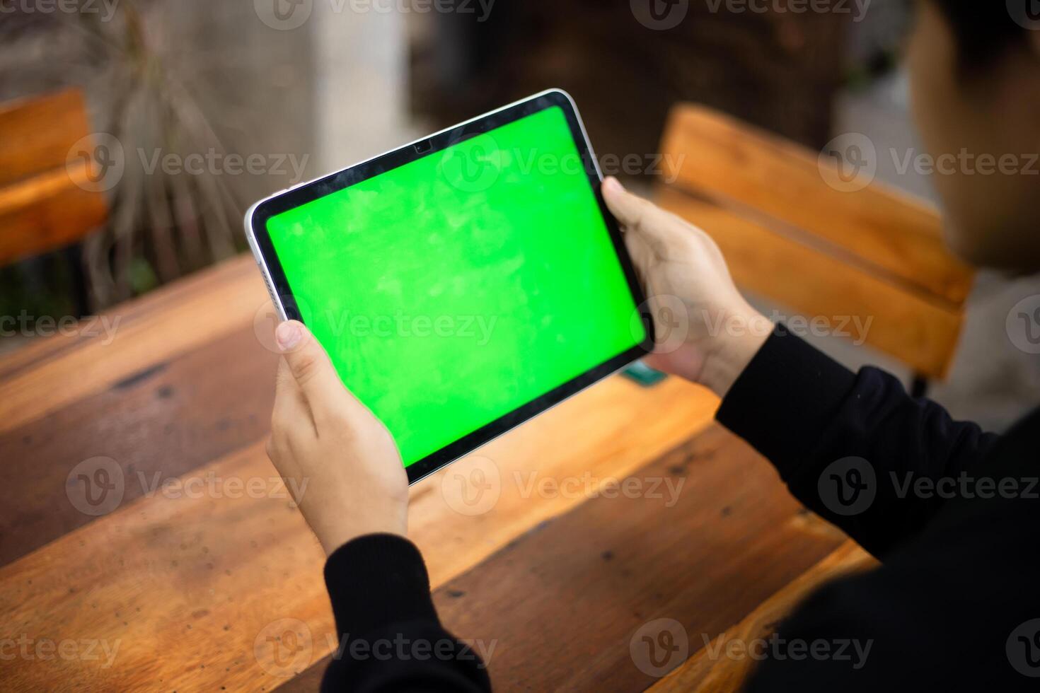 Mock up photo of a close up shot featuring a mans hand holding an iPad tablet with a green screen against the background of a wood cafe table