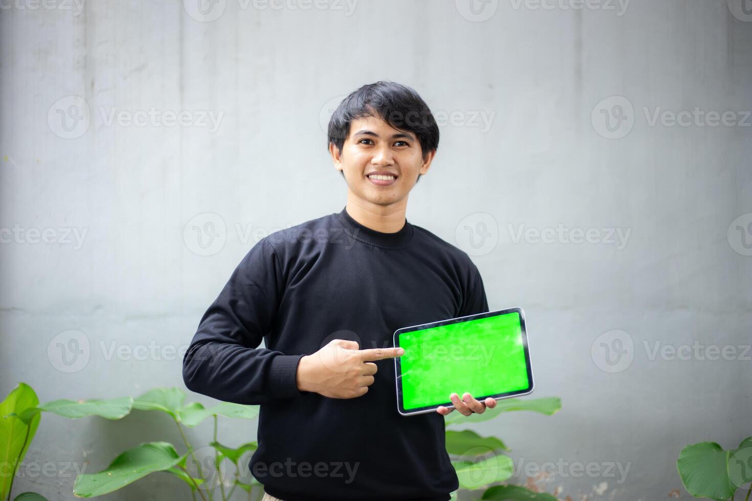 Young Asian man with a black sweater holding and pointing at an iPad tablet green screen mockup photo