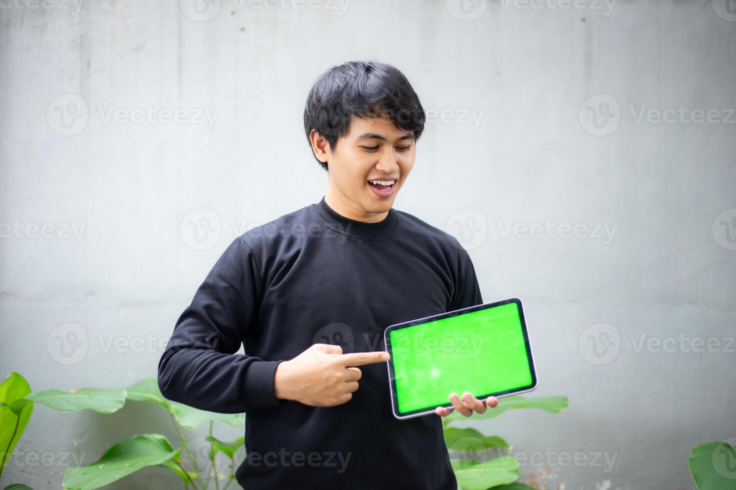 Young Asian man with a black sweater holding and pointing at an iPad tablet green screen mockup photo