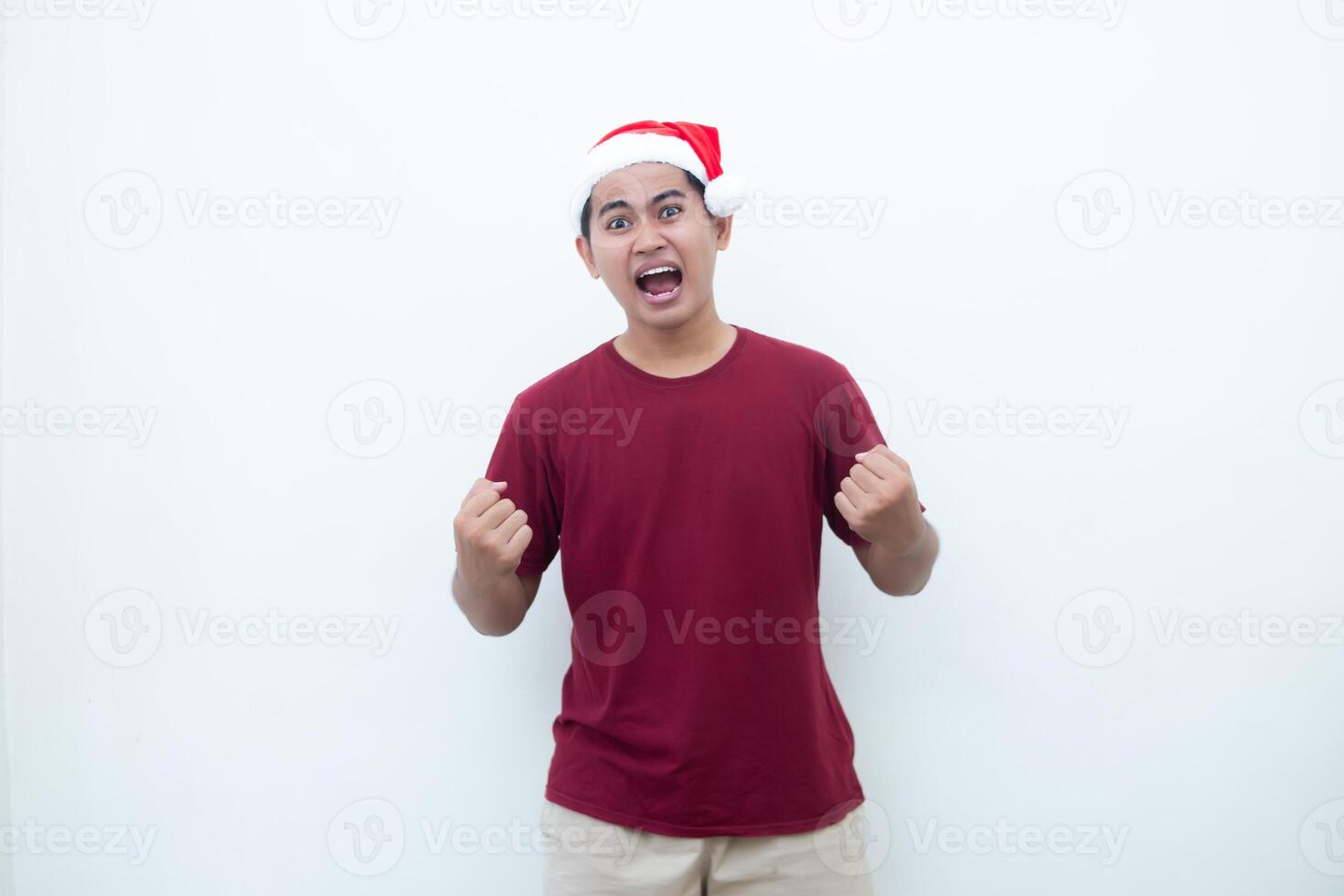 Young Asian man wearing a Santa Claus hat gripping both hands with enthusiasm and anger isolated by a white background for visual communication photo