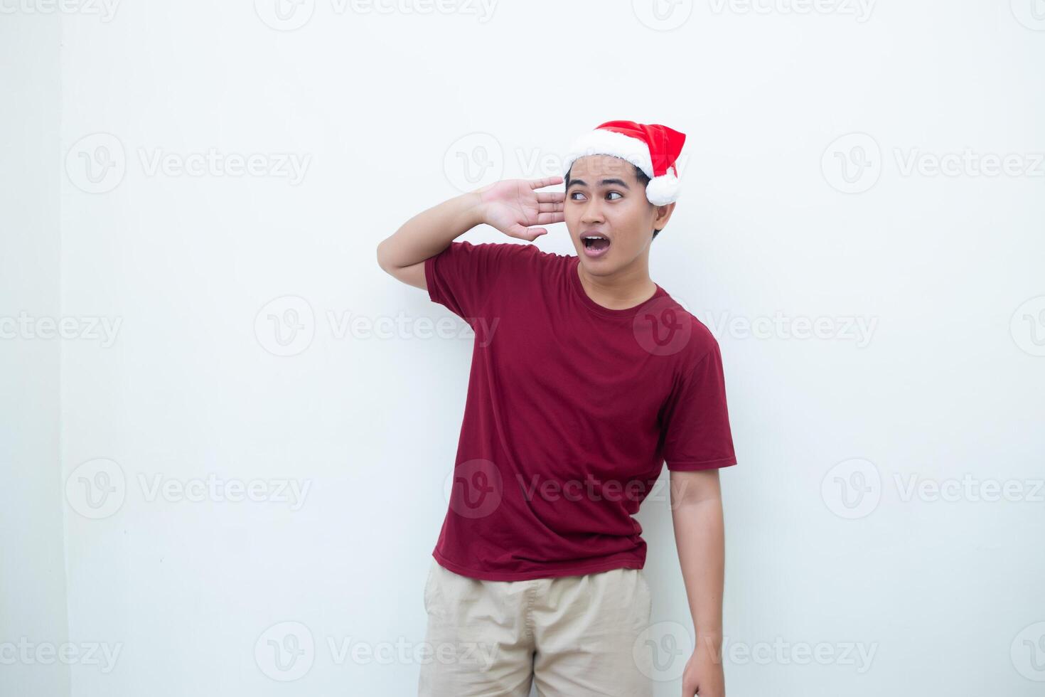 Young Asian man wearing a Santa Claus hat expressing listening with hand to ear isolated by a white background for visual communication photo