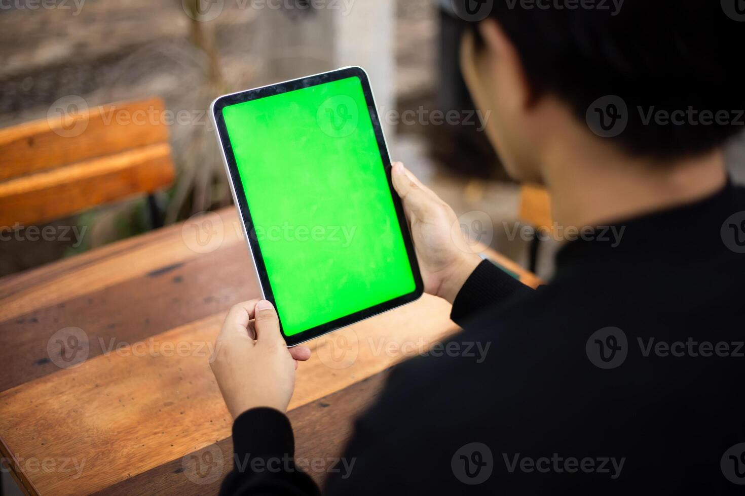 Mock up photo of a close up shot featuring a mans hand holding an iPad tablet with a green screen against the background of a wood cafe table