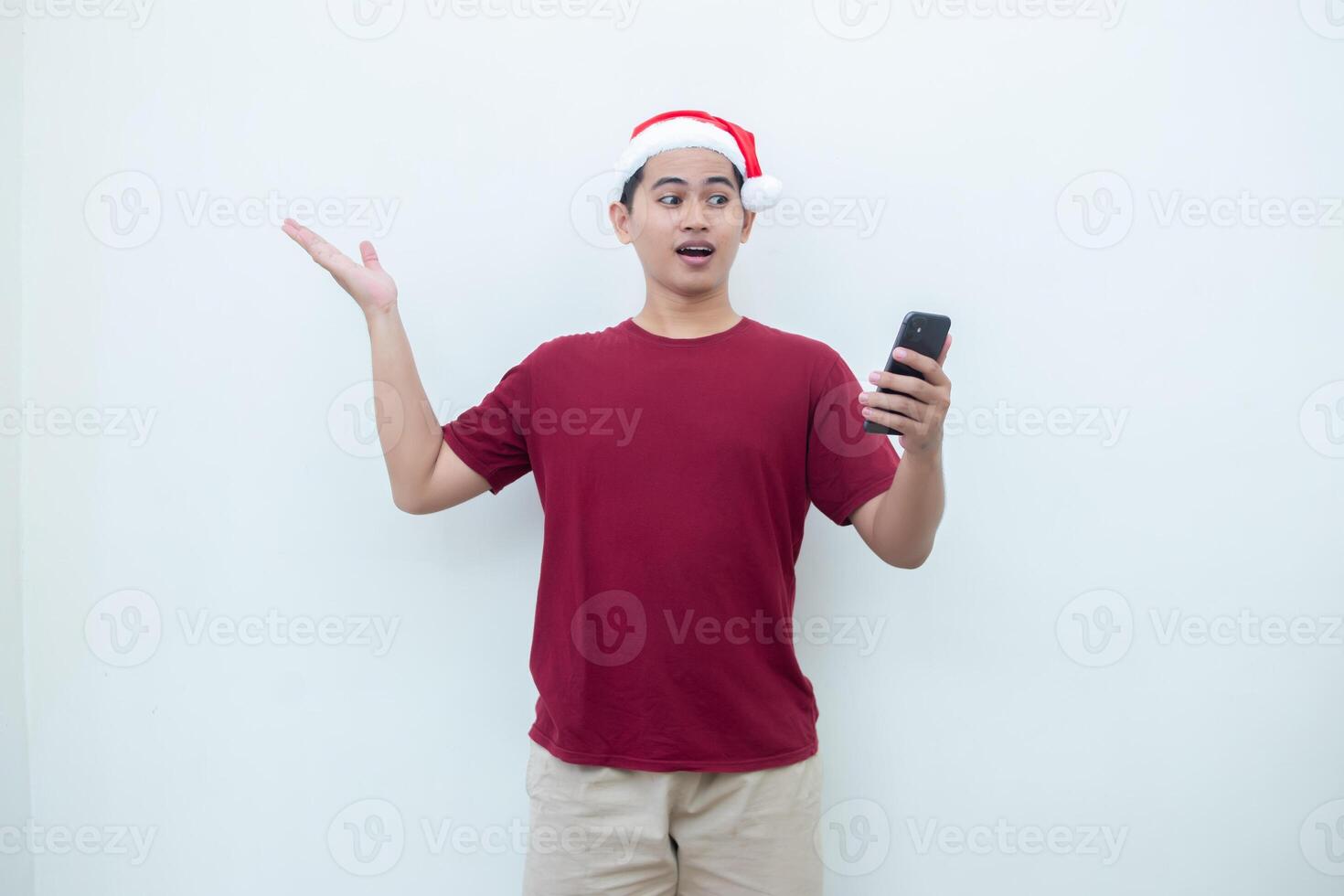 Young Asian man wearing a Santa Claus hat holding a smartphone and expressing a smile, surprise, and pointing to the side, isolated by a white background for visual communication photo
