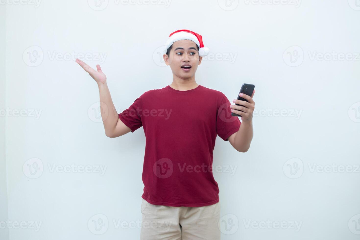 joven asiático hombre vistiendo un Papa Noel claus sombrero participación un teléfono inteligente y expresando un sonrisa, sorpresa, y señalando a el lado, aislado por un blanco antecedentes para visual comunicación foto