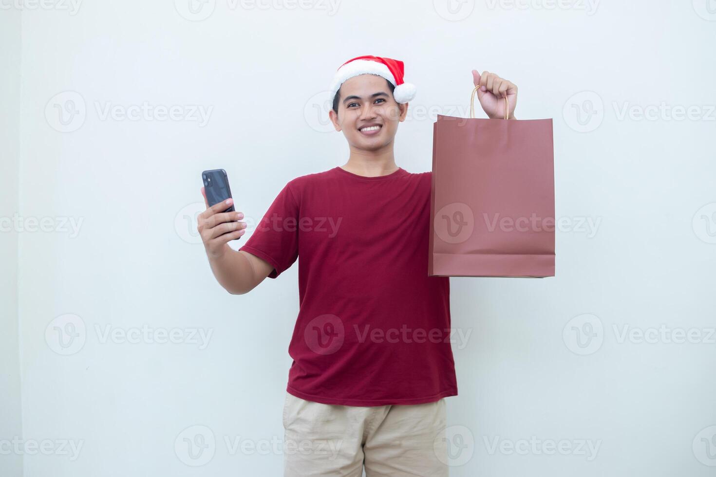 Young Asian man wearing a Santa Claus hat holding a smartphone and a shopping bag with expressions of smile, shock, and surprise, isolated against a white background for visual communication photo