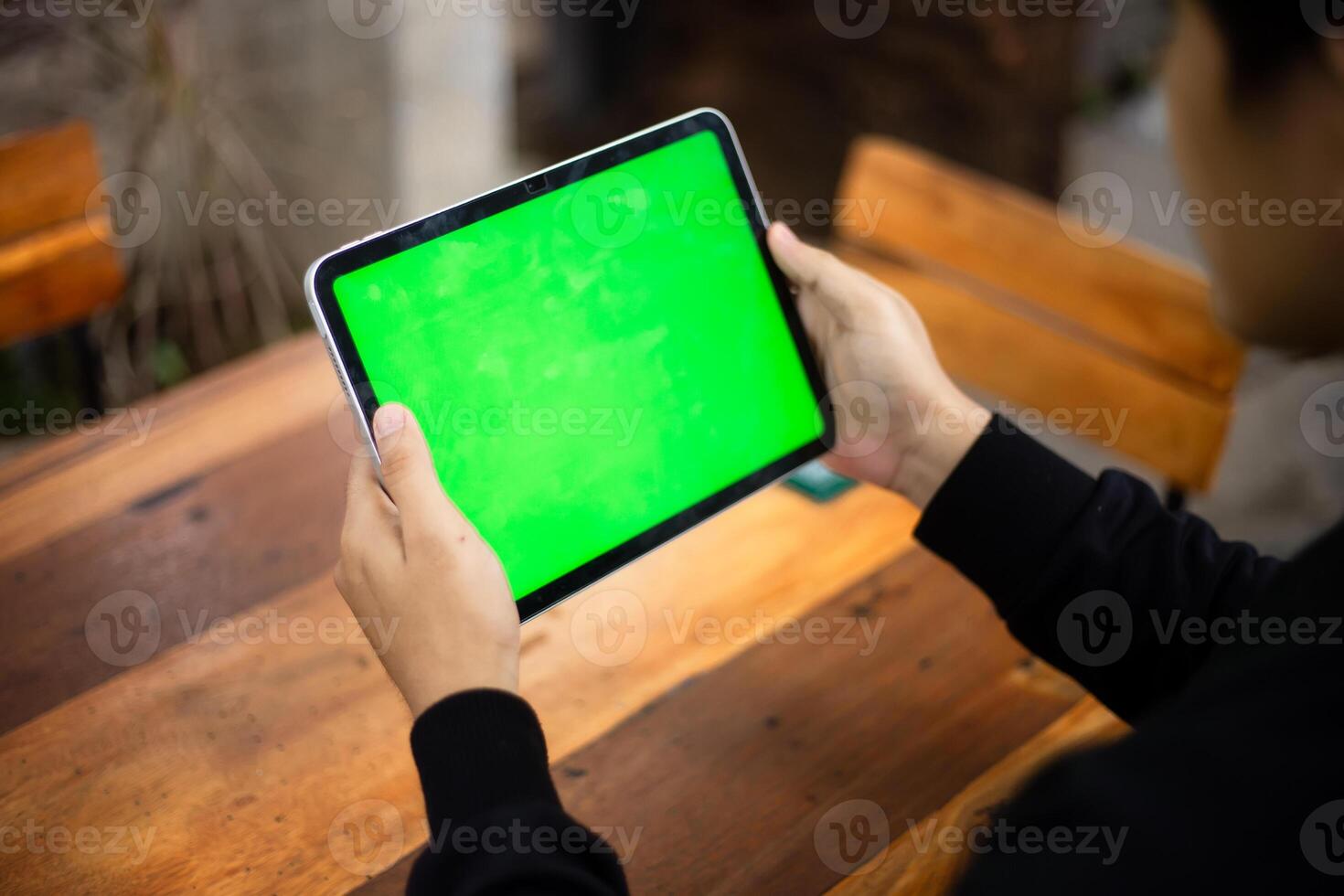 Mock up photo of a close up shot featuring a mans hand holding an iPad tablet with a green screen against the background of a wood cafe table