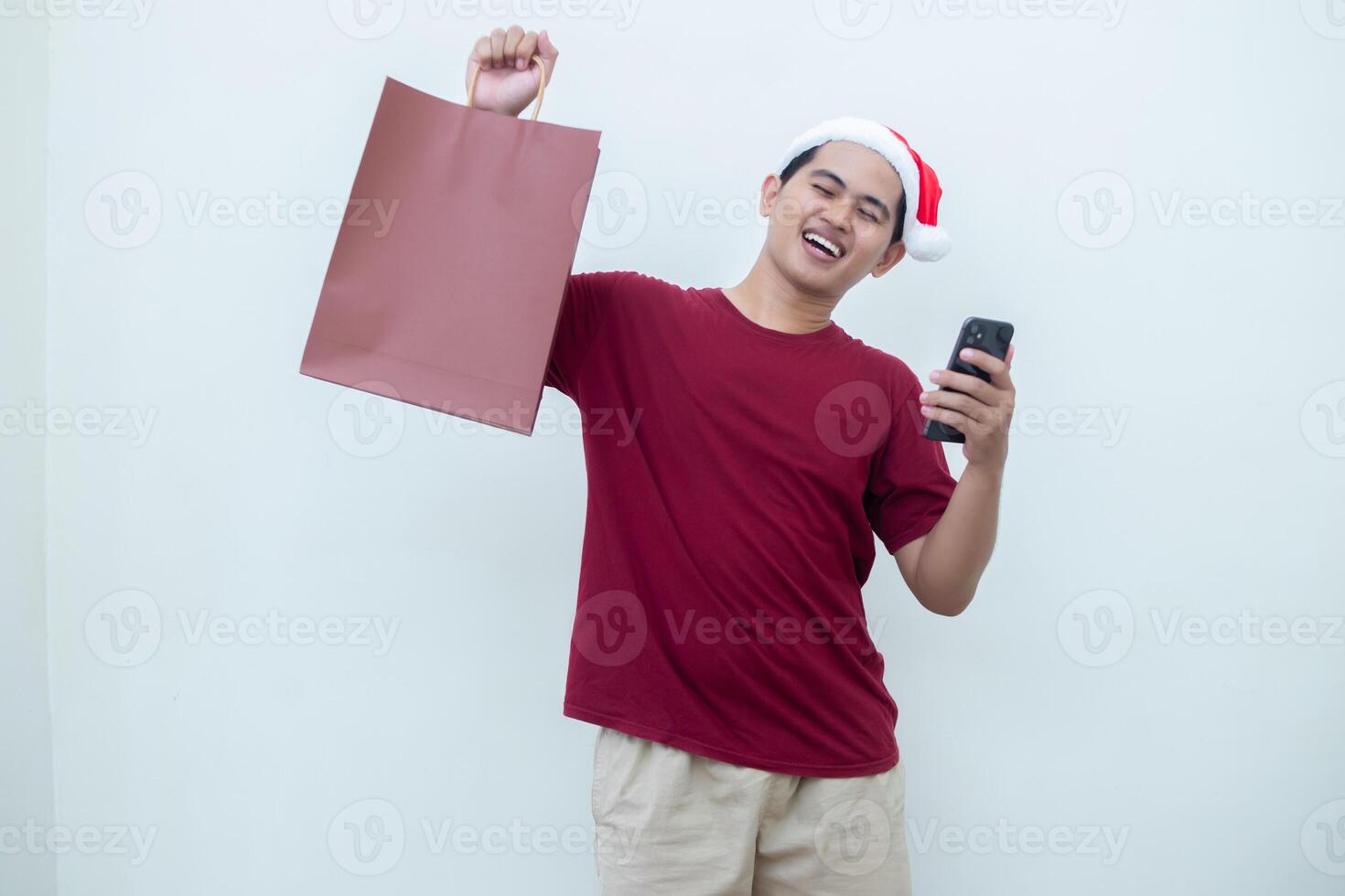 Young Asian man wearing a Santa Claus hat holding a smartphone and a shopping bag with expressions of smile, shock, and surprise, isolated against a white background for visual communication photo