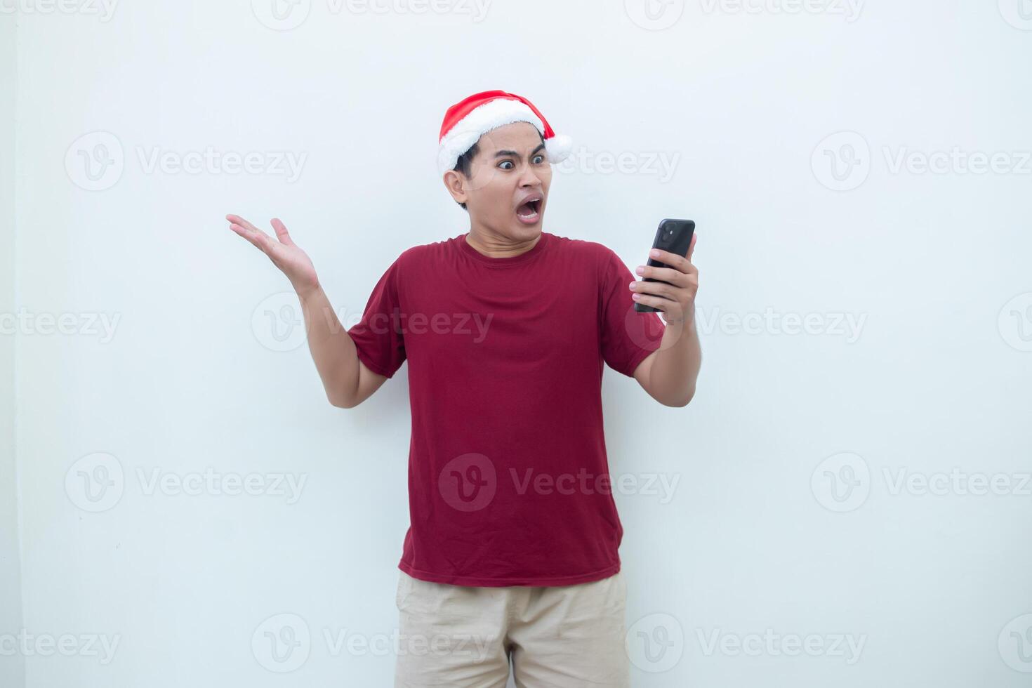 Young Asian man wearing a Santa Claus hat holding a smartphone and expressing a smile, surprise, and pointing to the side, isolated by a white background for visual communication photo