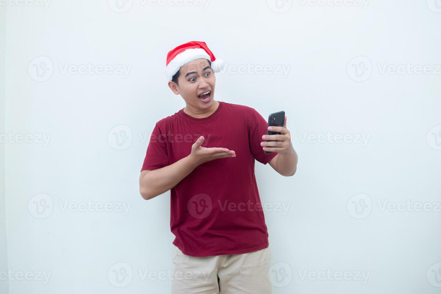 Young Asian man wearing a Santa Claus hat holding a smartphone and expressing a smile, surprise, and pointing to the side, isolated by a white background for visual communication photo