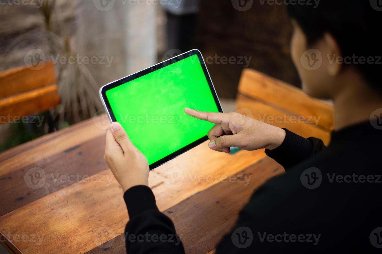 Mock up photo of a close up shot featuring a mans hand holding an iPad tablet with a green screen against the background of a wood cafe table