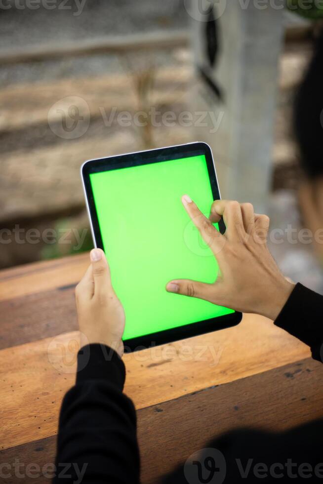 Mock up photo of a close up shot featuring a mans hand holding an iPad tablet with a green screen against the background of a wood cafe table