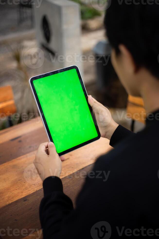 Mock up photo of a close up shot featuring a mans hand holding an iPad tablet with a green screen against the background of a wood cafe table